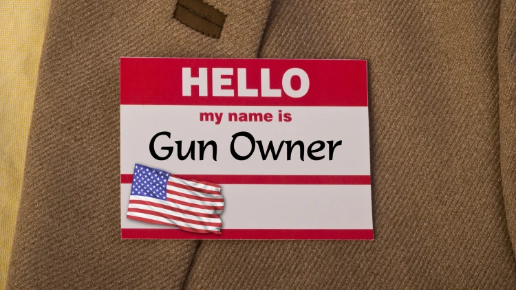 Close-up view of red and white name tag pinned to brown cloth, displaying text: 'Hello my name is gun owner'.