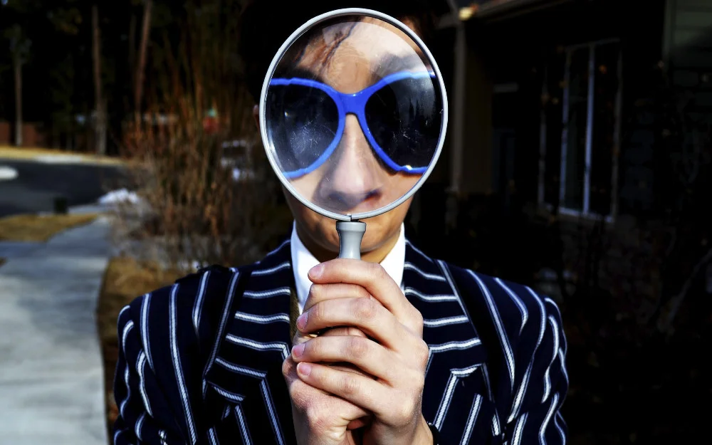 Man in striped suit holding a magnifying glass up to his face, wearing large blue sunglasses.