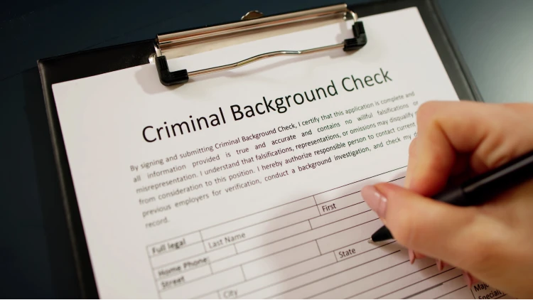 Close-up view of a criminal background check form on a clipboard, with a hand holding a pen writing on it.