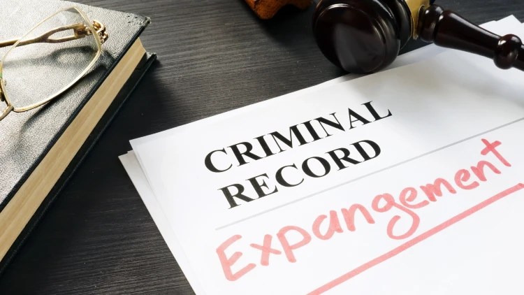 Close-up view of a book with eyeglasses on top, alongside a gavel and pieces of paper printed with the words 'criminal record,' with 'expungement' written below.