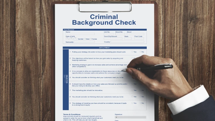 From a top-down perspective, a criminal background check form rests on a wooden table, with hands grasping a pen atop it.