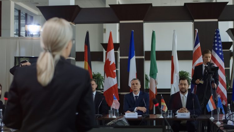 An image showing a meeting room with several international government representatives at a table, the flags of various countries behind them, and a woman's back to the camera in the foreground