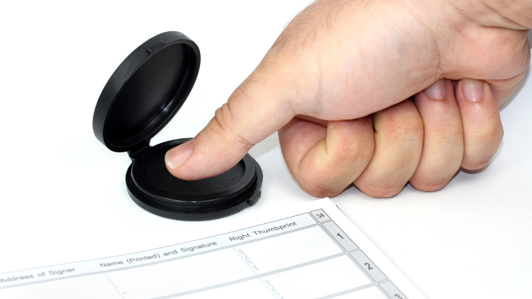 A person's right hand presses its thumb onto an ink pad to take a fingerprint next to a form requiring a fingerprint.