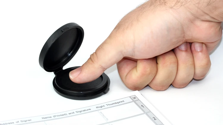 A person's right hand presses its thumb onto an ink pad to take a fingerprint next to a form requiring a fingerprint.
