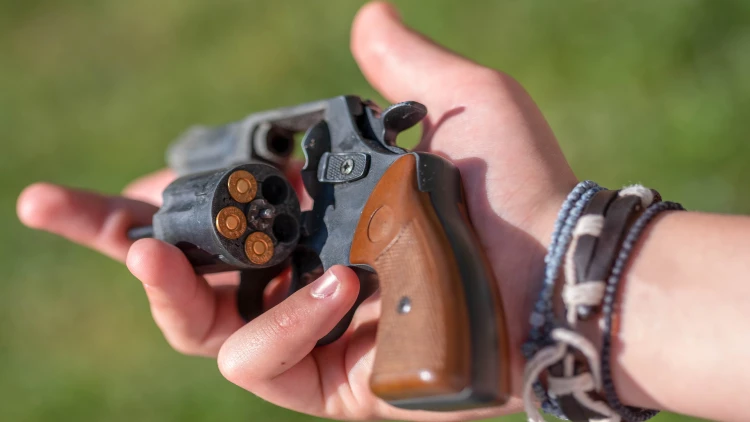 Close-up of a hand holding a revolver with the cylinder open, showing four bullets loaded. The person, possibly practicing concealed carry, is wearing bracelets on their wrist. Background is blurred green foliage.