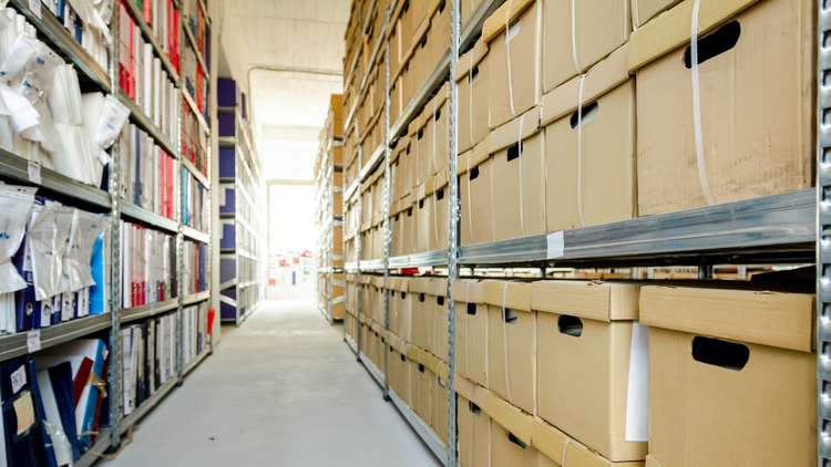 An aisle between tall shelves filled with cardboard boxes and files in a public records archive room.