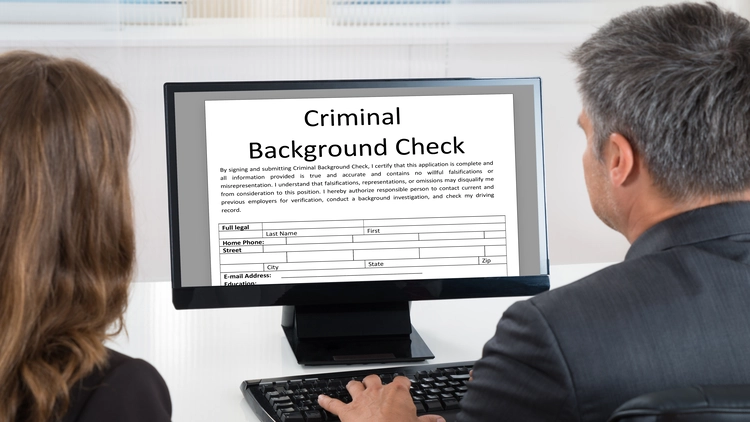 Two professionals sit at a desk, viewing a criminal background check form on a computer screen.