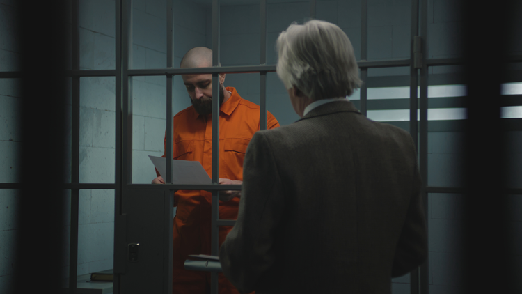 A lawyer stands outside a jail cell, speaking with a bearded inmate in an orange jumpsuit who is reading a document, in a dimly lit prison.