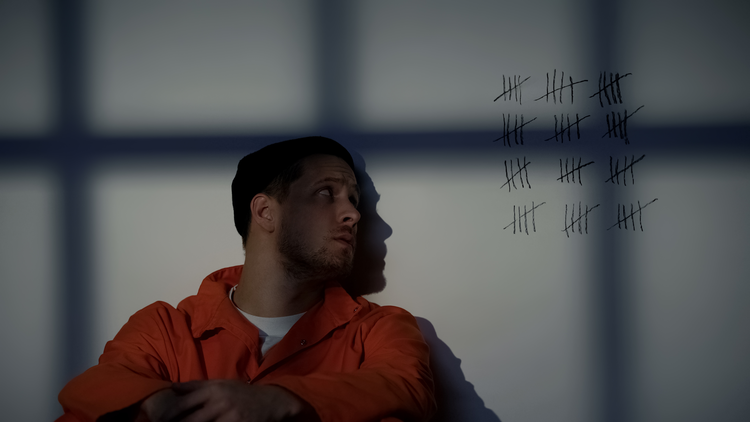 A male inmate in an orange jumpsuit sits against a wall with tally marks, indicating days spent in prison, drawn beside him, while shadows from bars fall across the scene.