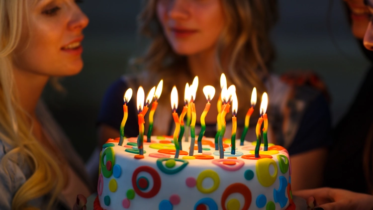 A woman blowing out lit candles on a colorful birthday cake in a darkened room with her friends.