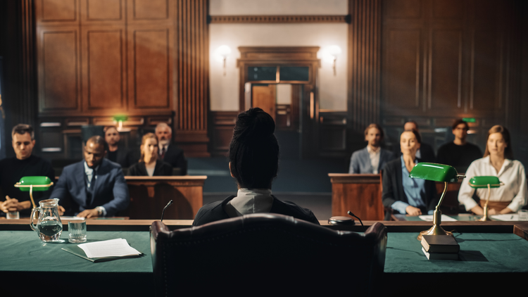 A courtroom scene with a person sitting at a desk facing a jury and audience.