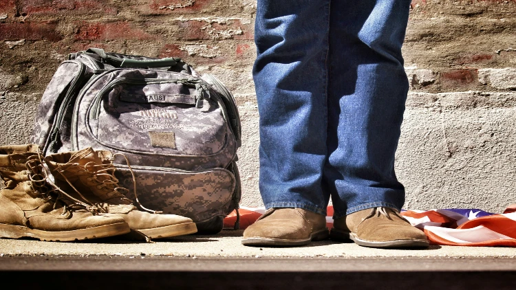 Close up image of a man's legs and feet in casual wear besides a military bag and boots and the U.S. flag