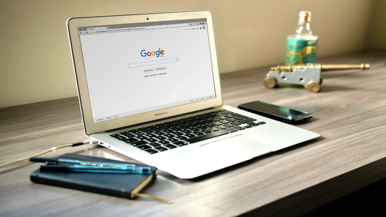 Image of Google search engine open in a laptop screen on top of a desk with notebook, pens, and phone