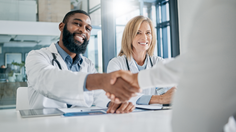 Two healthcare professionals shaking hands across a desk, representing a successful job interview or employment agreement.