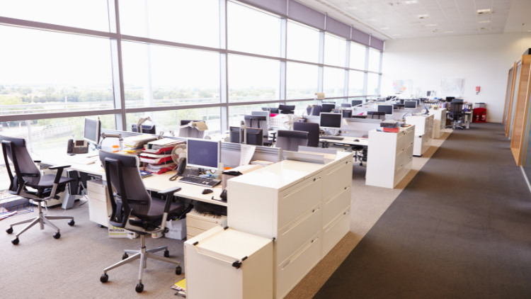 A modern open-plan office with rows of desks and chairs, each with computers and office supplies, sits eerily still. Large windows provide natural light, but the space feels empty, as if recently abandoned.