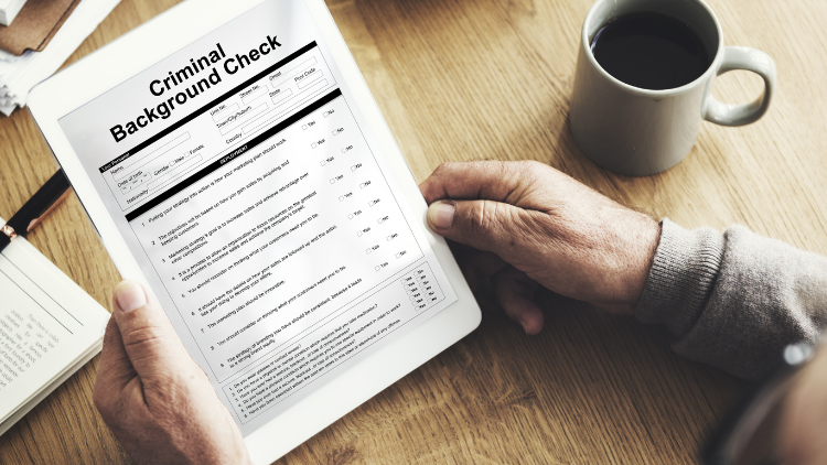From a top-down perspective, a criminal background check form rests on a wooden table, with hands grasping a pen atop it.