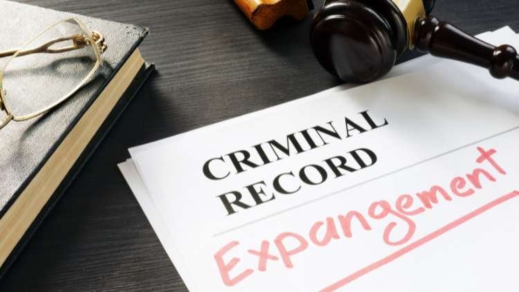 Close-up view of a book with eyeglasses on top, alongside a gavel and pieces of paper printed with the words 'criminal record,' with 'expungement' written below.