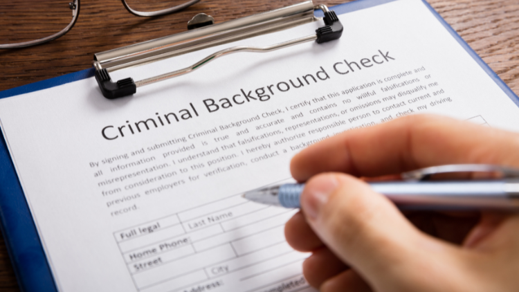 Close-up view of a criminal background check form on a clipboard, with a hand holding a pen writing on it.