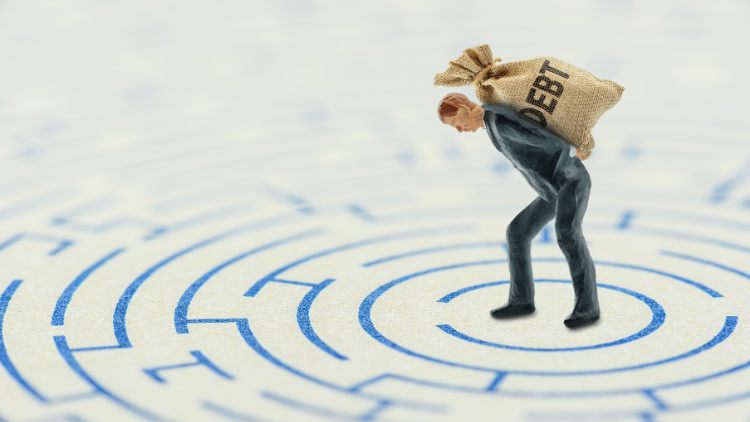 A toy man standing in the middle of a drawn maze, carrying a woven sack with the word 'debt' written on it.