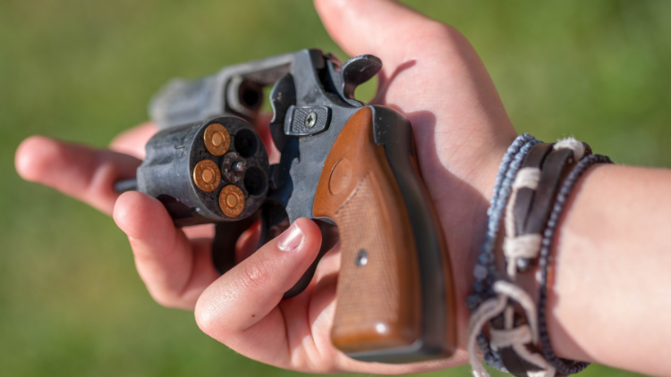 Close-up of a hand holding a revolver with the cylinder open, showing four bullets loaded. The person, possibly practicing concealed carry, is wearing bracelets on their wrist. Background is blurred green foliage.