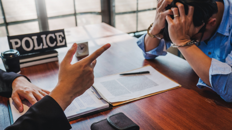 Police officer's hand pointing towards man in handcuffs on wooden table.