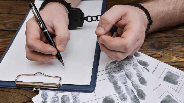 A man's hand in handcuffs holding a pen on a blank white paper clipped on a clipboard.