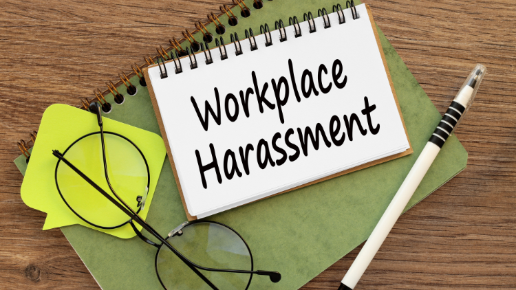 A notebook with Workplace Harassment written on it, next to glasses, a pen, and sticky notes on a wooden surface.