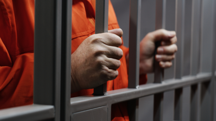 Close-up view of an elderly prisoner in an orange uniform holding metal bars, standing in a prison cell.