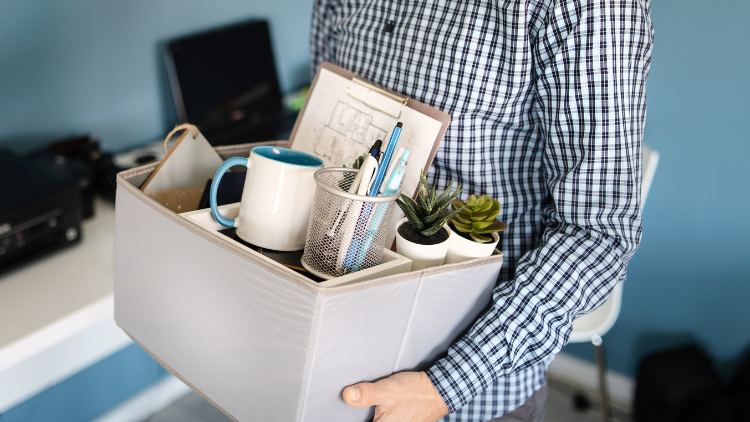 A man wearing a checkered shirt holds a box containing office supplies, including a mug, pens, a planner, a potted plant, and a pencil holder, suggesting he might have just been fired.
