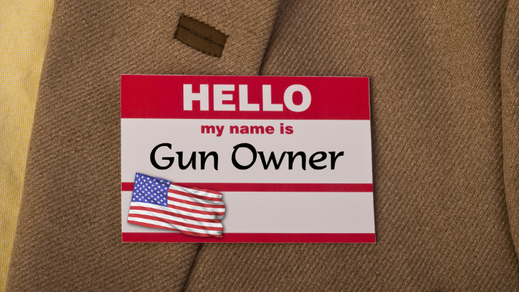 Close-up view of red and white name tag pinned to brown cloth, displaying text: 'Hello my name is gun owner'.